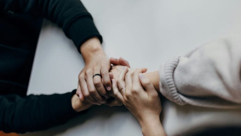 Two people holding hands in a supportive gesture, with one wearing a black ring.