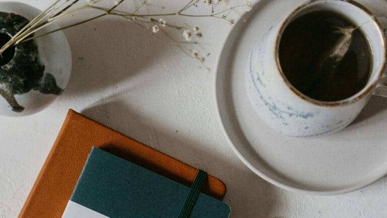 Coffee cup on a saucer beside two notebooks and dried flowers on a white tabletop